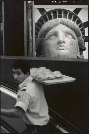 A Very American Picture: a waiter carrying cheeseburgers with a poster of the Statue of Liberty in the background. “It becomes a collage in one frame through [Rauschenberg’s] camera lens,” explains Sean Corcoran, senior curator of prints and photographs at the Museum of the City of New York.