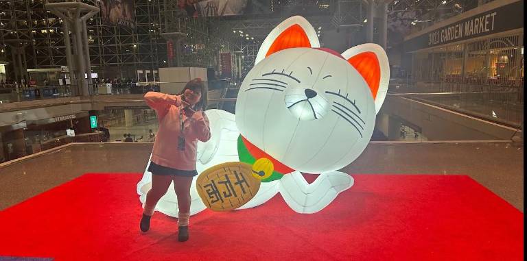 An Anime NYC attendee cosplaying Miss Aisa poses beside a giant display of Turbo Granny in luck cat form from “Dandadan.”
